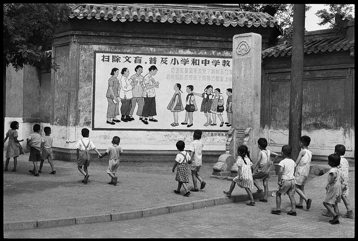 《孩童正從一幅寫著「&hellip;媽媽們都是上學去&hellip;」宣傳標語的看板前經過，北京，1958年7月》